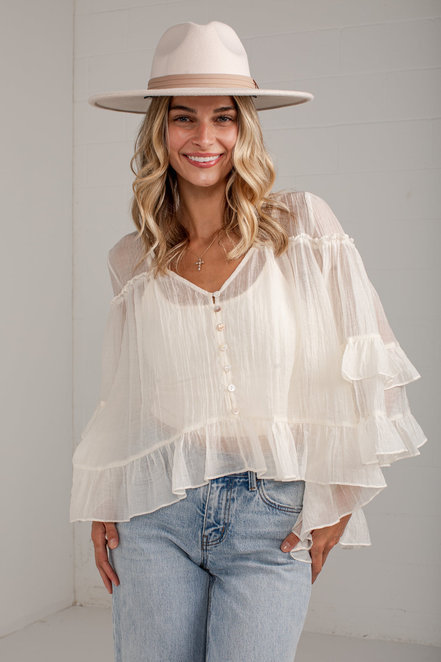 A woman wearing a sheer white tiered blouse with ruffled sleeves, light blue jeans, and a beige wide-brim hat, standing against a white paneled wall.