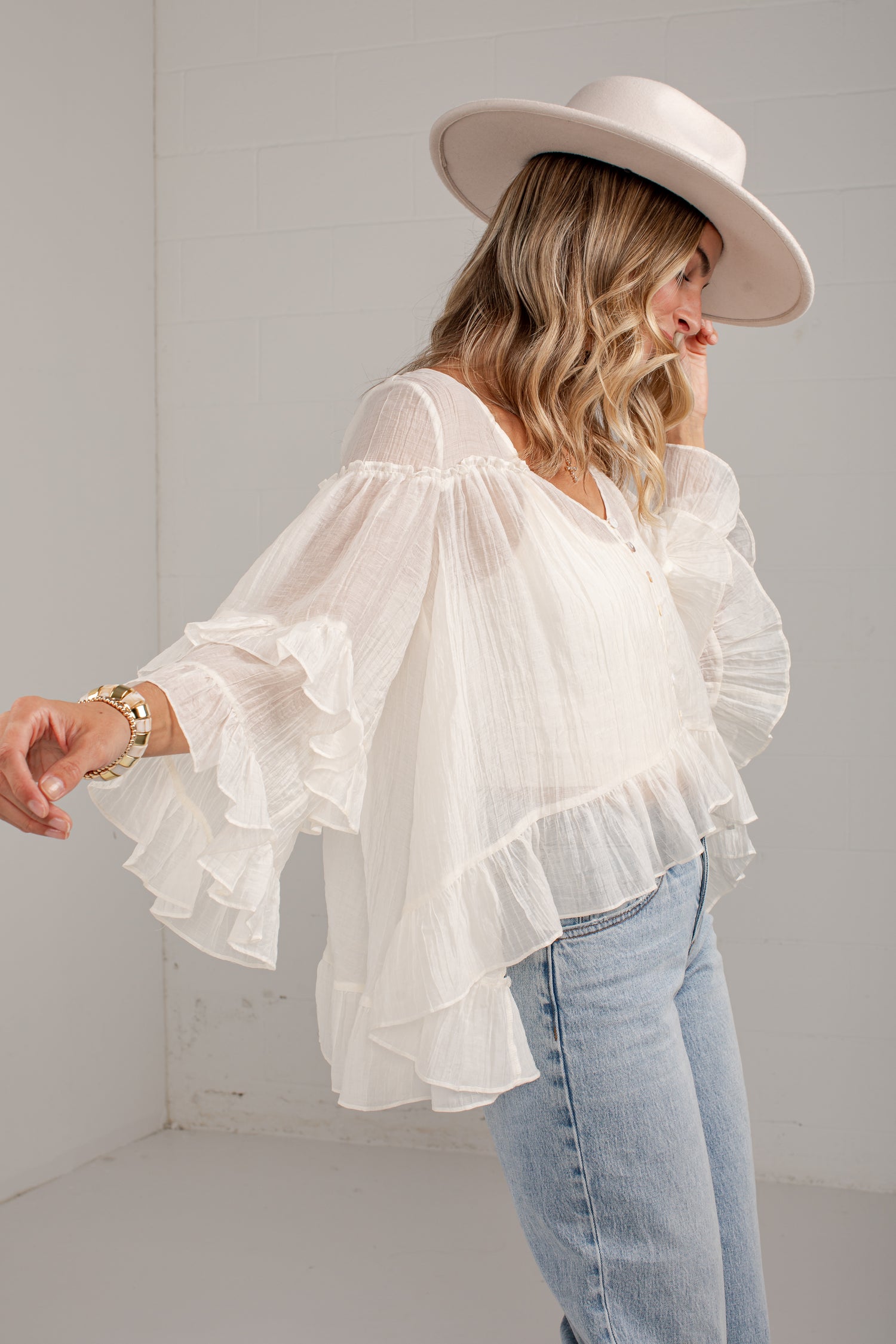 A woman wearing a white ruffled off-the-shoulder blouse with long bell sleeves, paired with light blue jeans and a wide-brimmed cream hat, standing in a bright white room.