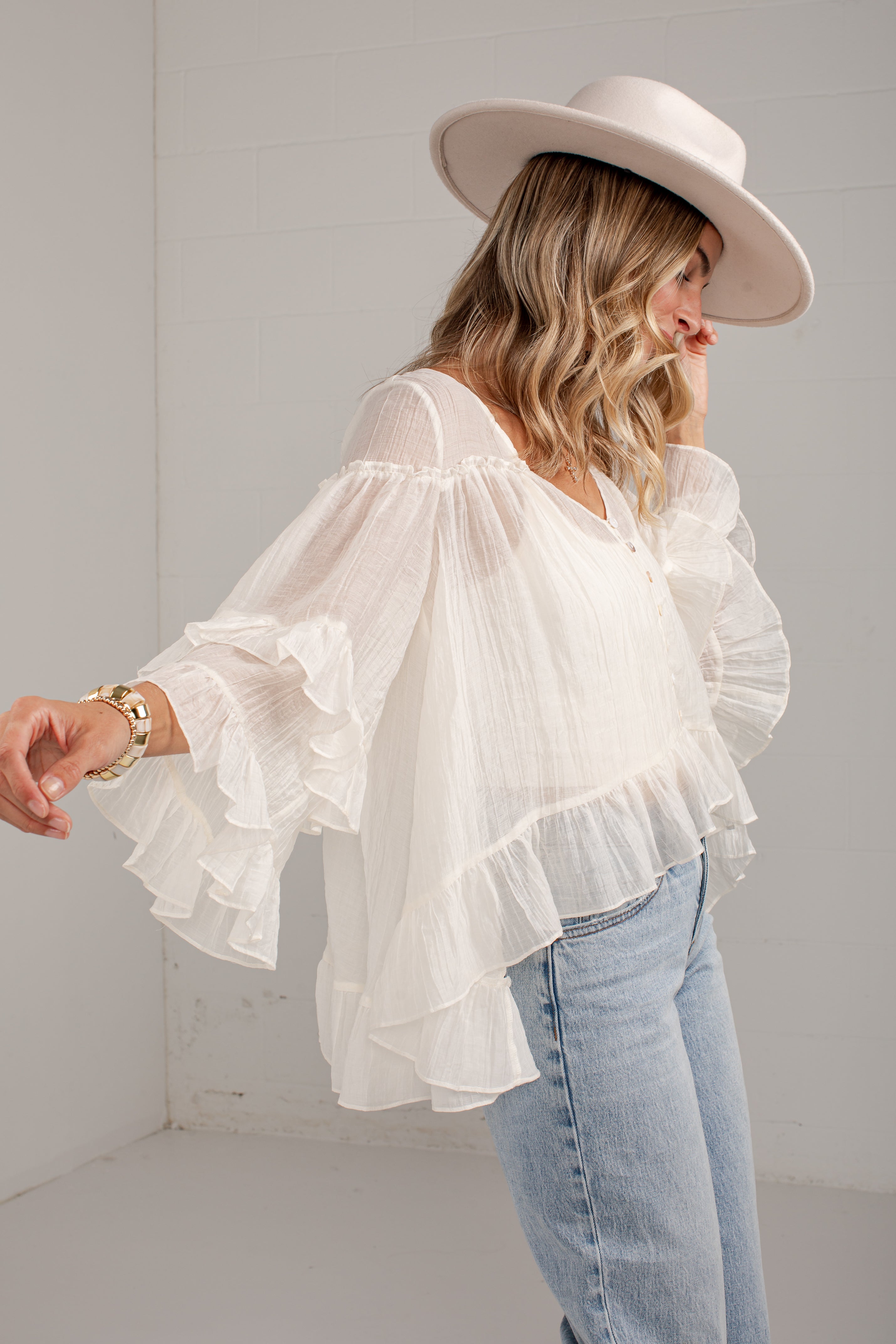 A woman wearing a white ruffled off-the-shoulder blouse with long bell sleeves, paired with light blue jeans and a wide-brimmed cream hat, standing in a bright white room.