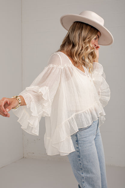 A woman wearing a white ruffled off-the-shoulder blouse with long bell sleeves, paired with light blue jeans and a wide-brimmed cream hat, standing in a bright white room.