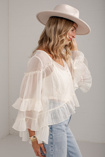 A woman wearing a white tiered ruffle blouse with sheer long sleeves, paired with light blue jeans and a wide-brimmed cream hat, standing against a white wall.