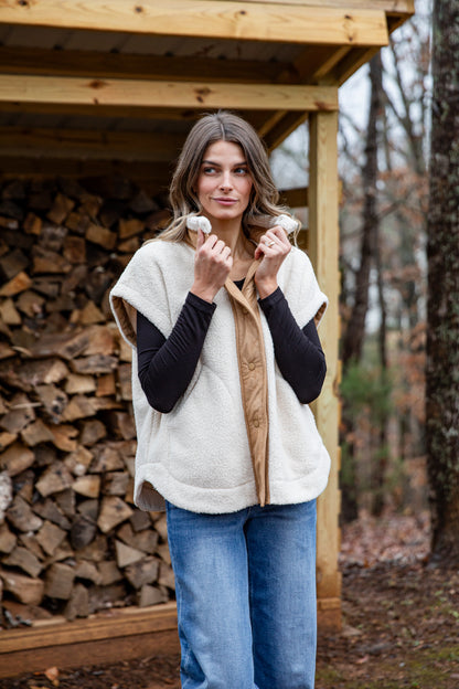 Woman wearing a white vest over a black long-sleeve shirt, standing in front of stacked firewood.