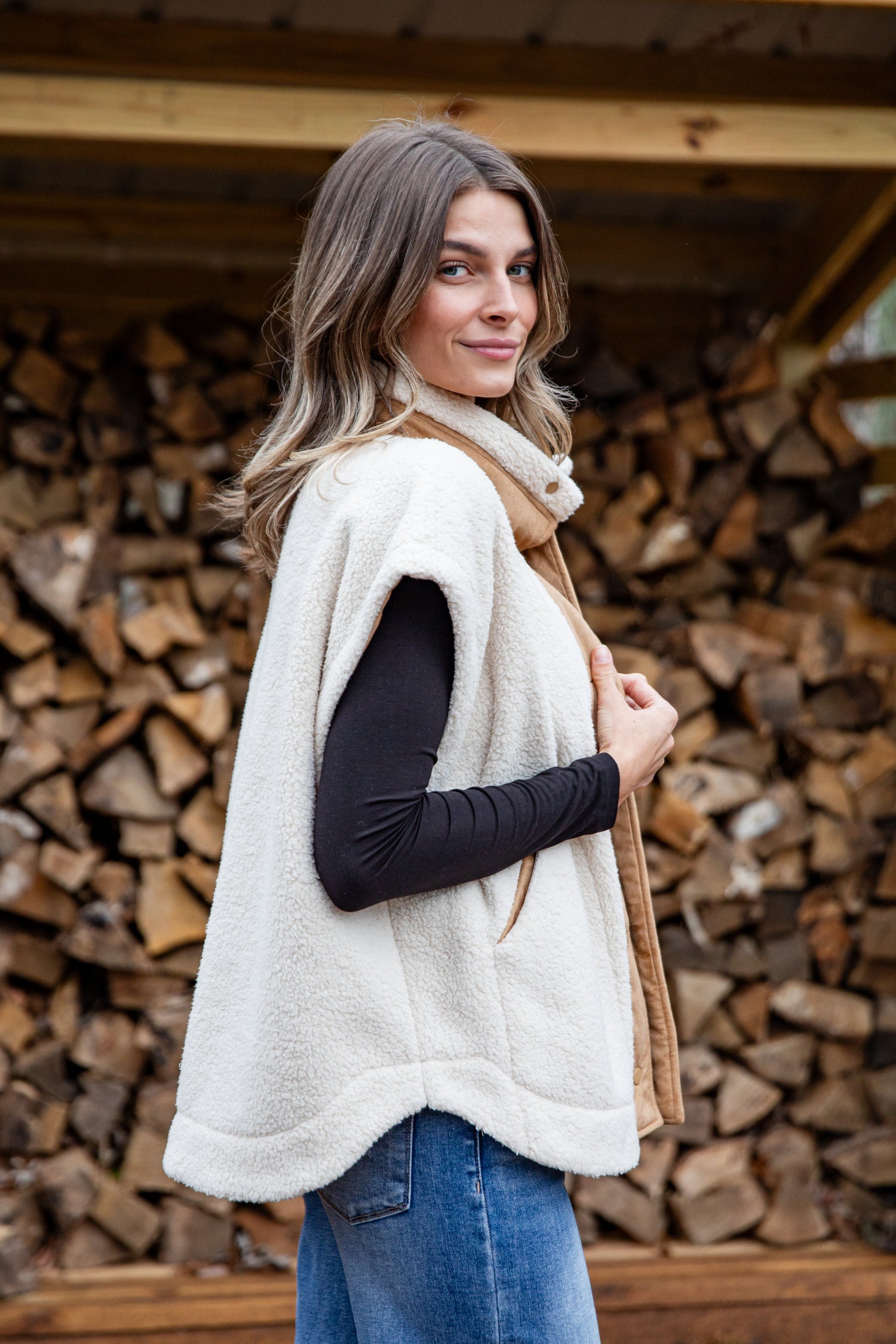 Woman wearing a white vest over a black long-sleeve shirt with a beige scarf, standing in front of stacked firewood.