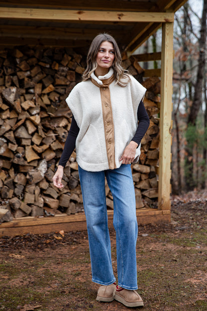 Woman wearing a white vest over a brown shirt with blue jeans, standing in front of a wooden structure with stacked firewood.