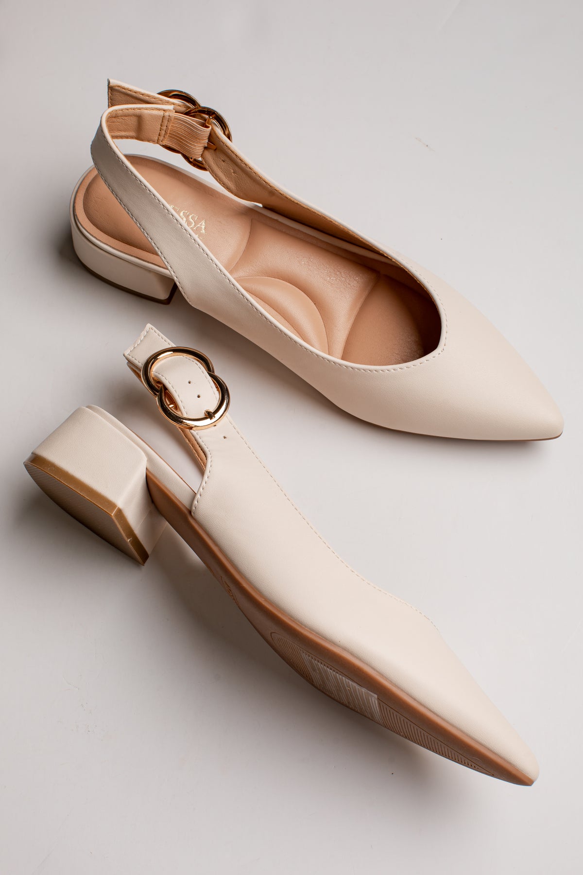 A pair of pointed-toe slingback flats in a warm cream color, featuring a low block heel and a decorative gold-tone buckle detail on the strap, displayed on a light gray background.