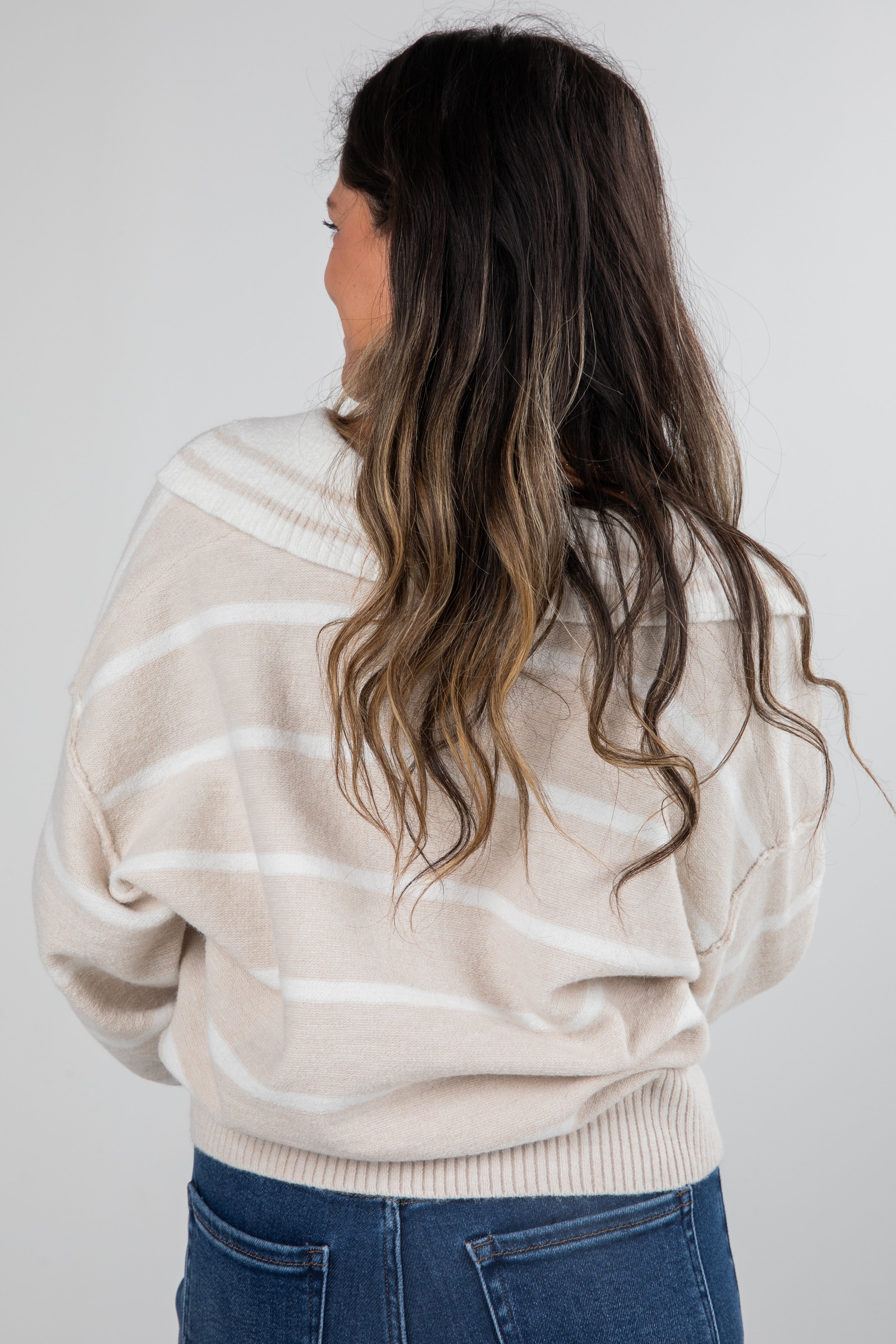 Person wearing a beige sweater with white stripes and blue jeans against a plain background