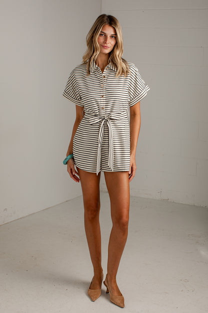 A woman models a short-sleeve collared romper with thin black and off-white horizontal stripes, featuring a button-up front and a tie waist. She is standing in a minimalist studio with white walls and a light gray floor.