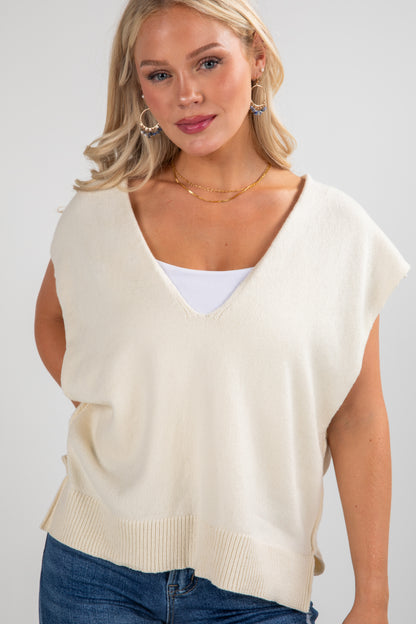 Woman wearing a beige sleeveless sweater over a white top with blue jeans on a plain background