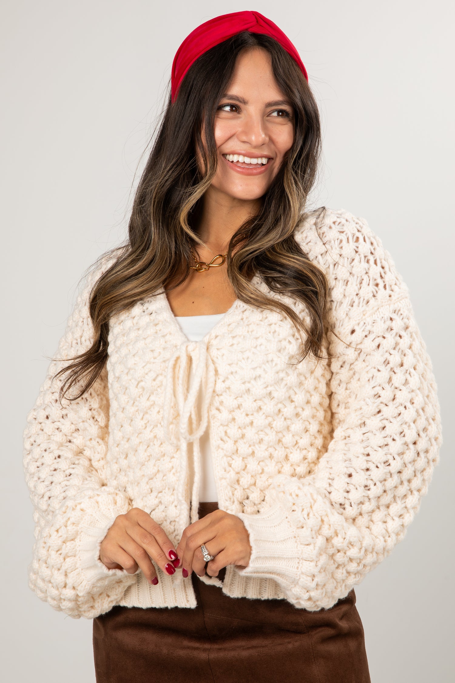 Woman wearing a cream chunky knit cardigan over a white top with a red headband on a plain background
