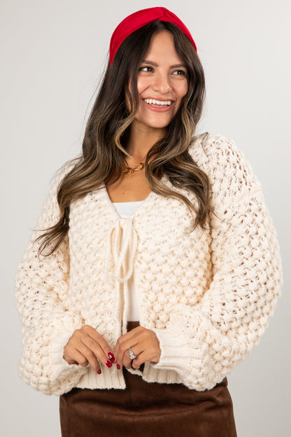 Woman wearing a cream chunky knit cardigan over a white top with a red headband on a plain background