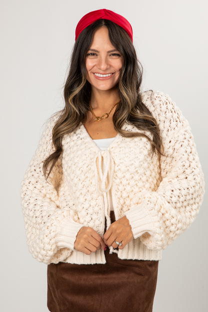 Woman wearing a cream chunky knit cardigan, brown pants, and a red headband on a white background