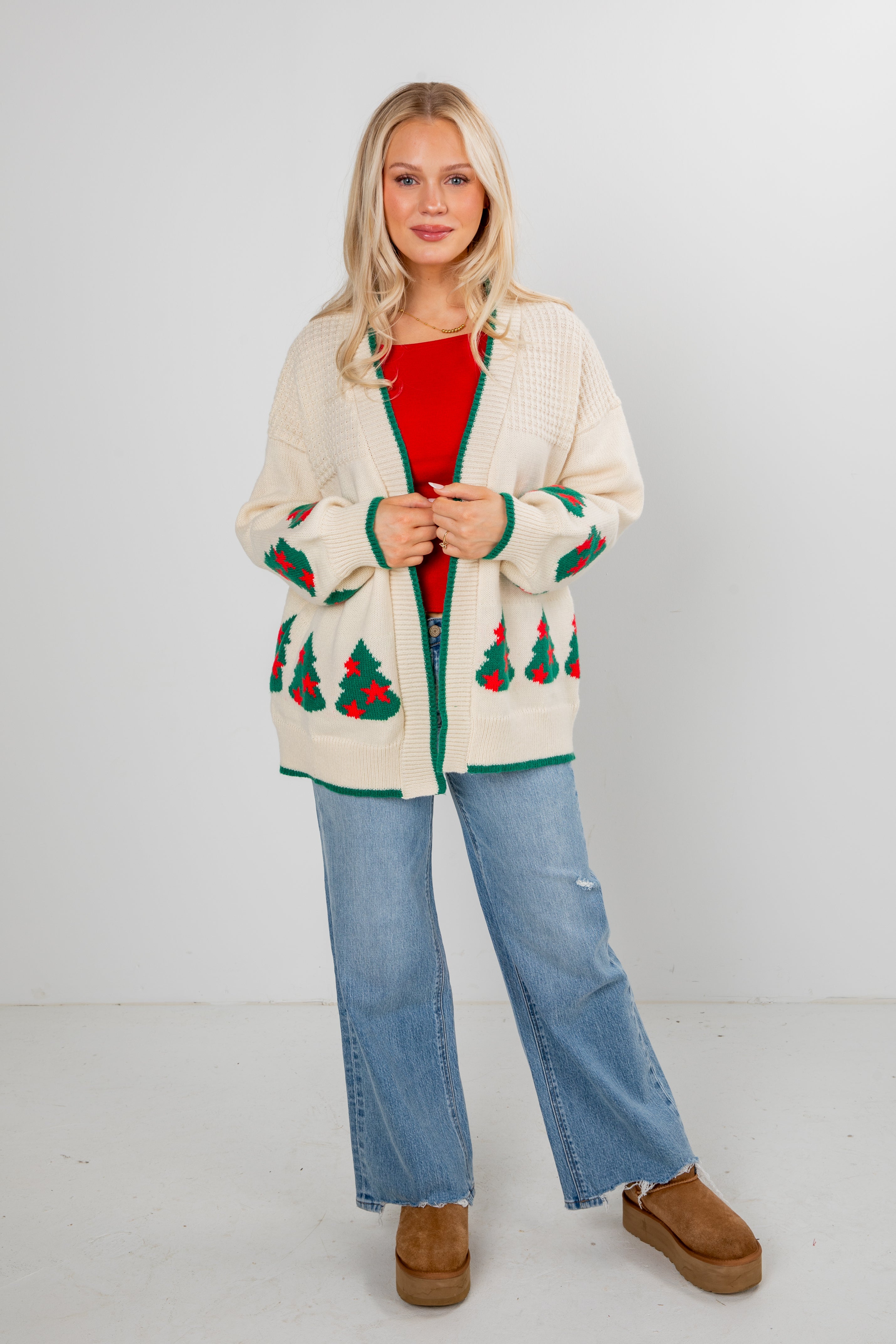 Woman wearing a cream cardigan with Christmas tree patterns, red shirt, blue jeans, and brown boots on a white background.
