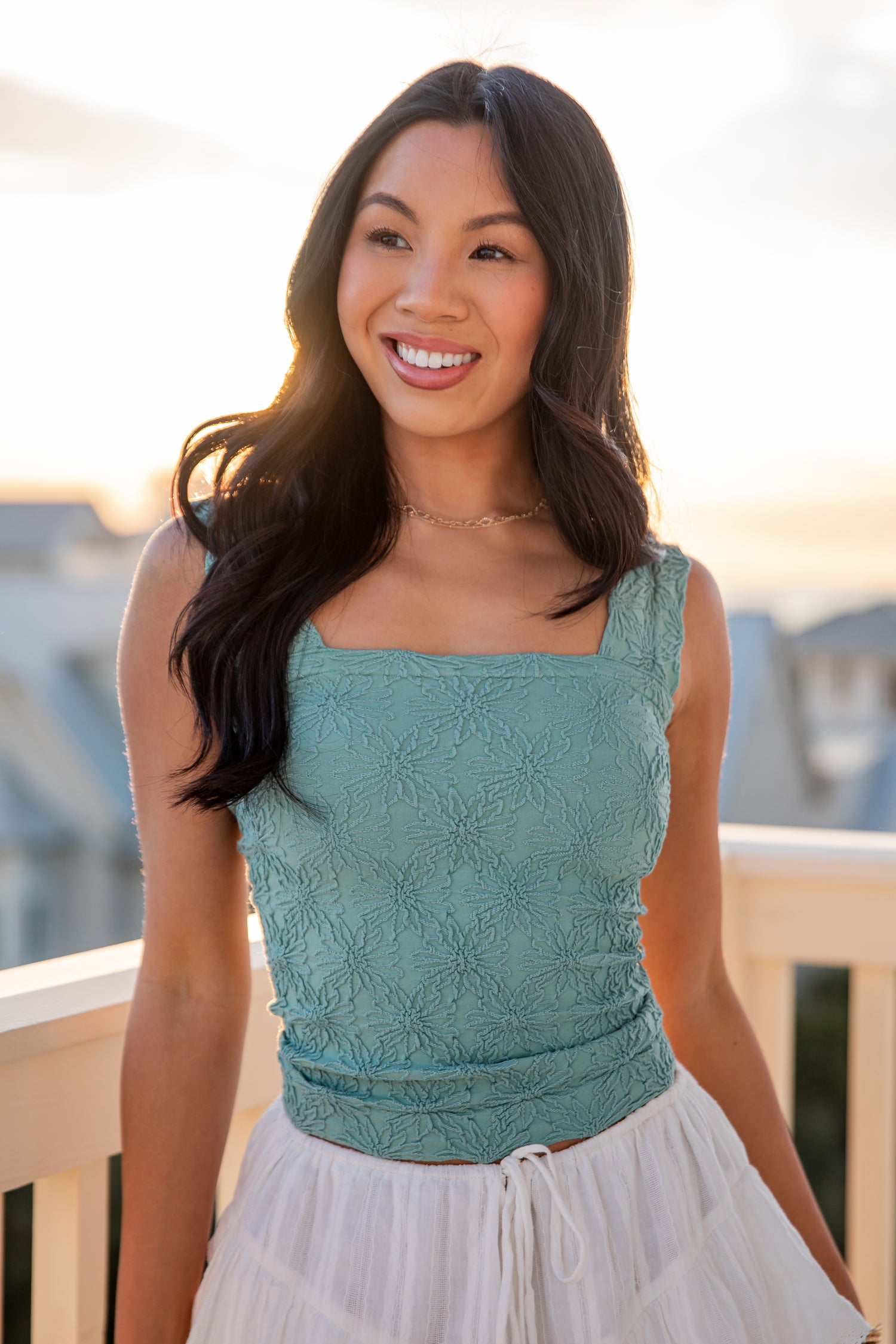 Woman wearing a teal lace top and white skirt on a balcony with a blurred background