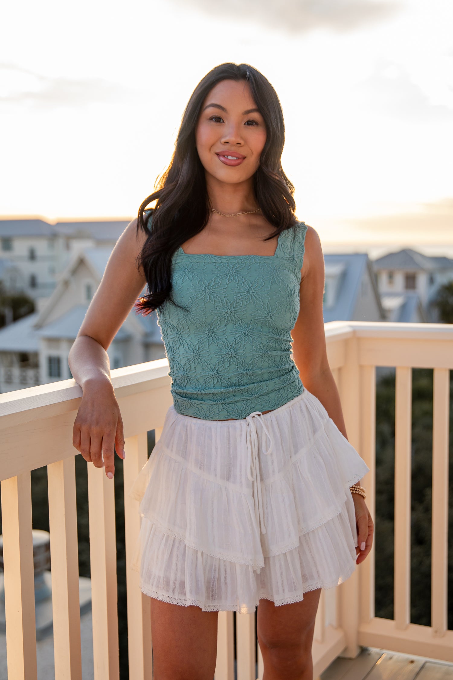 Woman in a teal top and white skirt standing on a balcony with a cityscape background