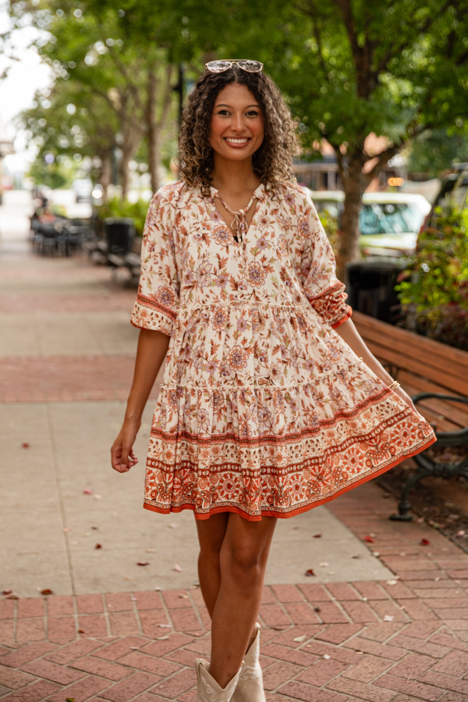 Woman in a floral dress standing on a sidewalk with trees and benches in the background