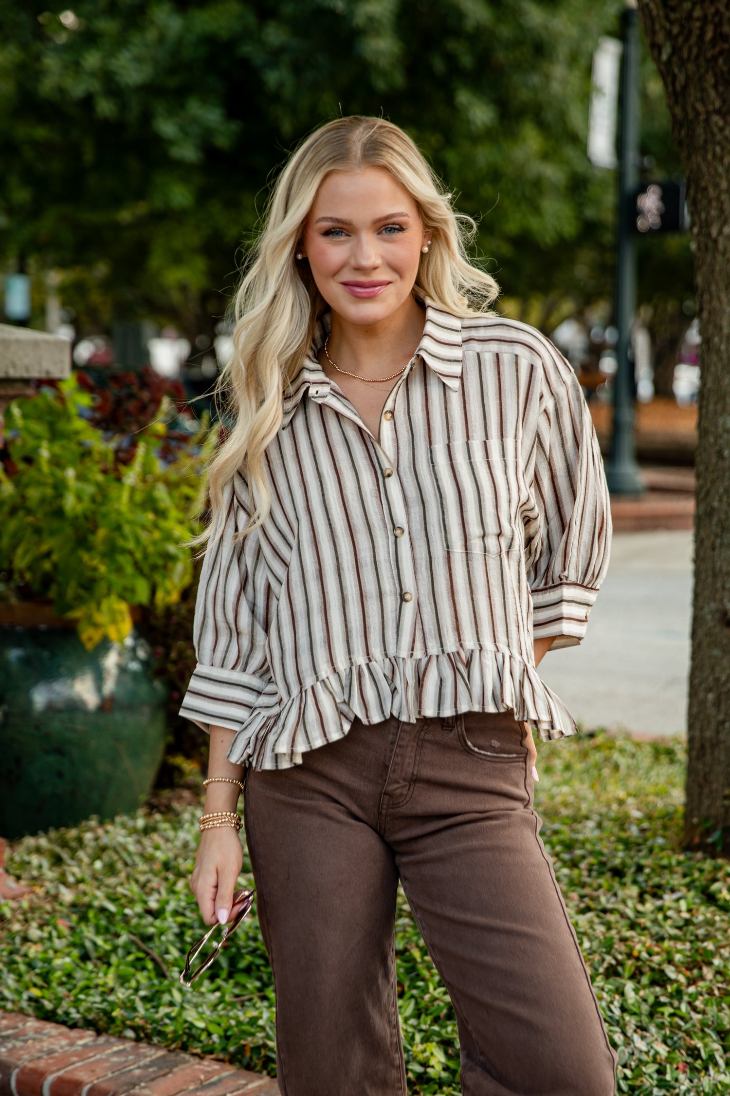 Woman wearing a striped shirt and brown pants standing outdoors with greenery in the background