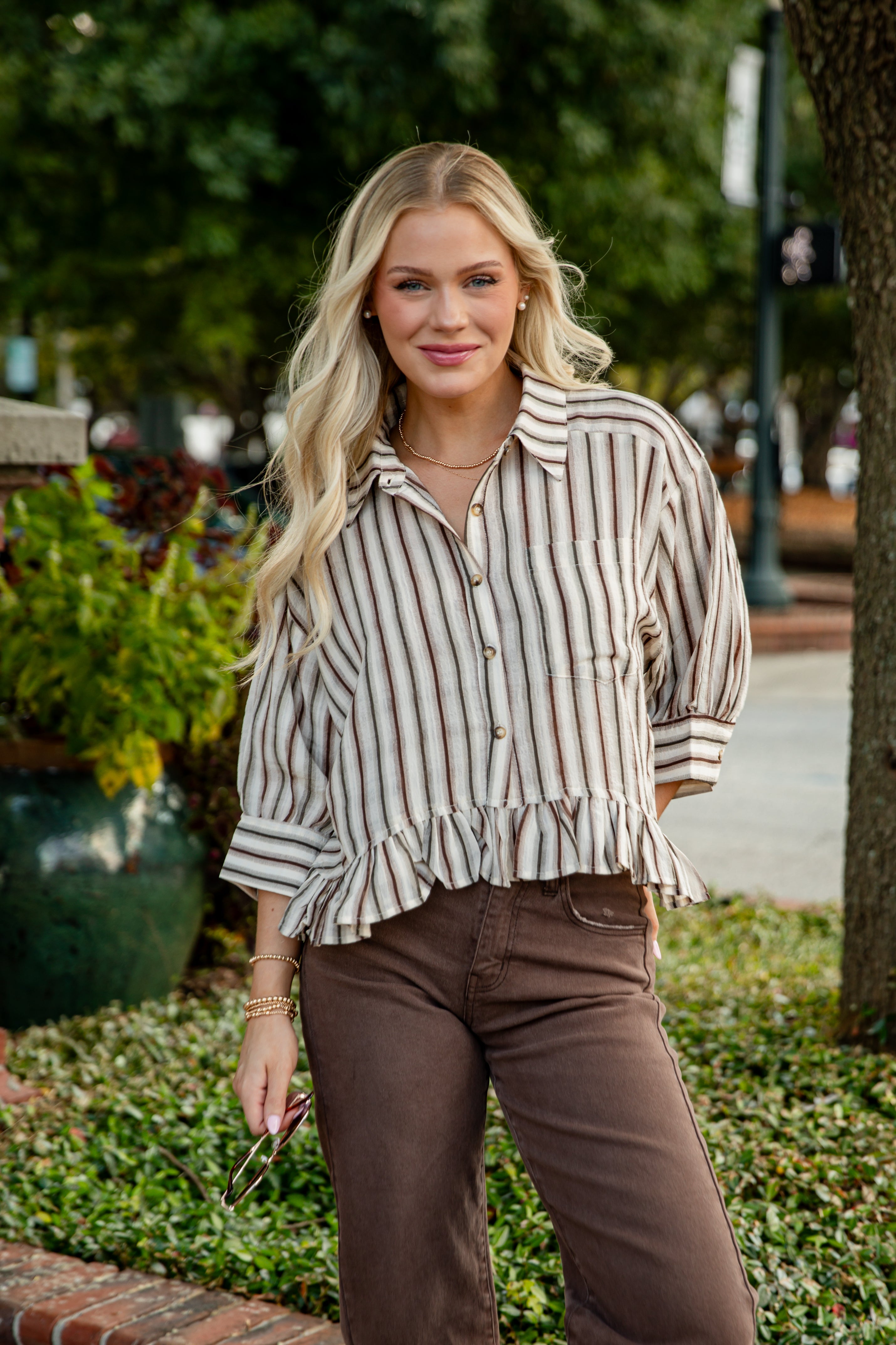 Woman wearing a striped shirt and brown pants standing outdoors with greenery in the background