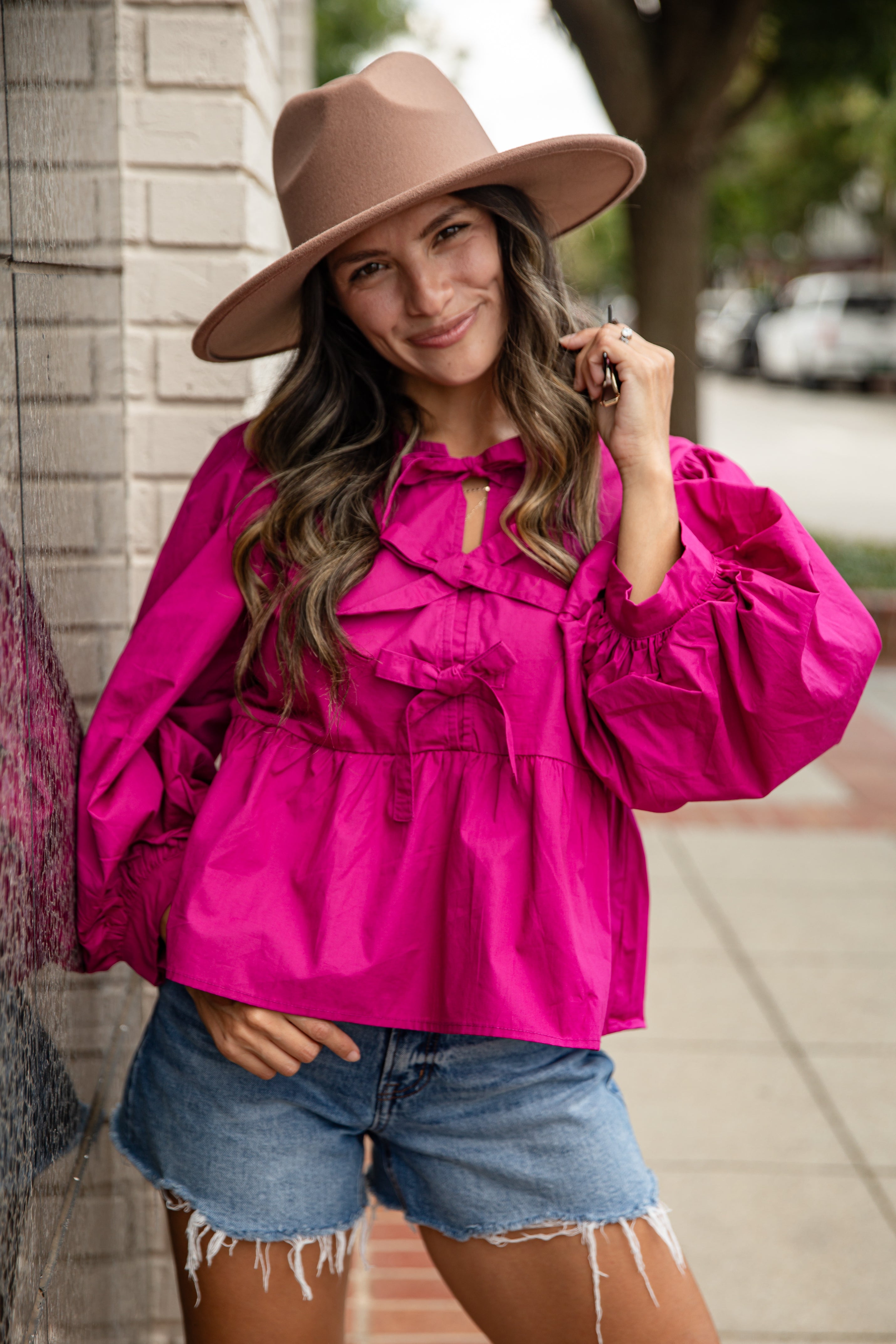 Woman wearing a bright pink blouse and denim shorts with a brown hat on a sidewalk.