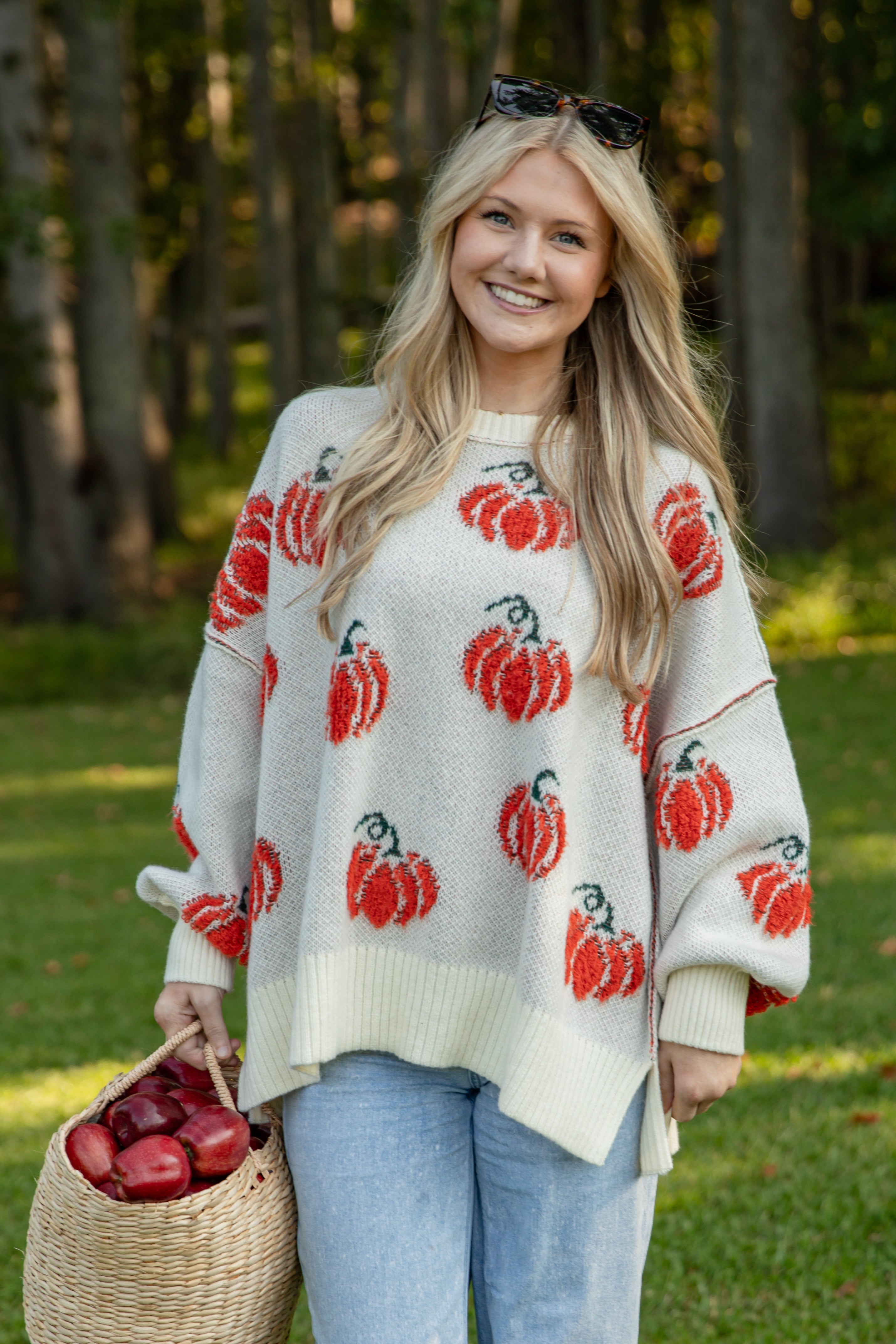 Woman wearing a cream-colored sweater with red pumpkin patterns, holding a basket of apples in a forest setting.