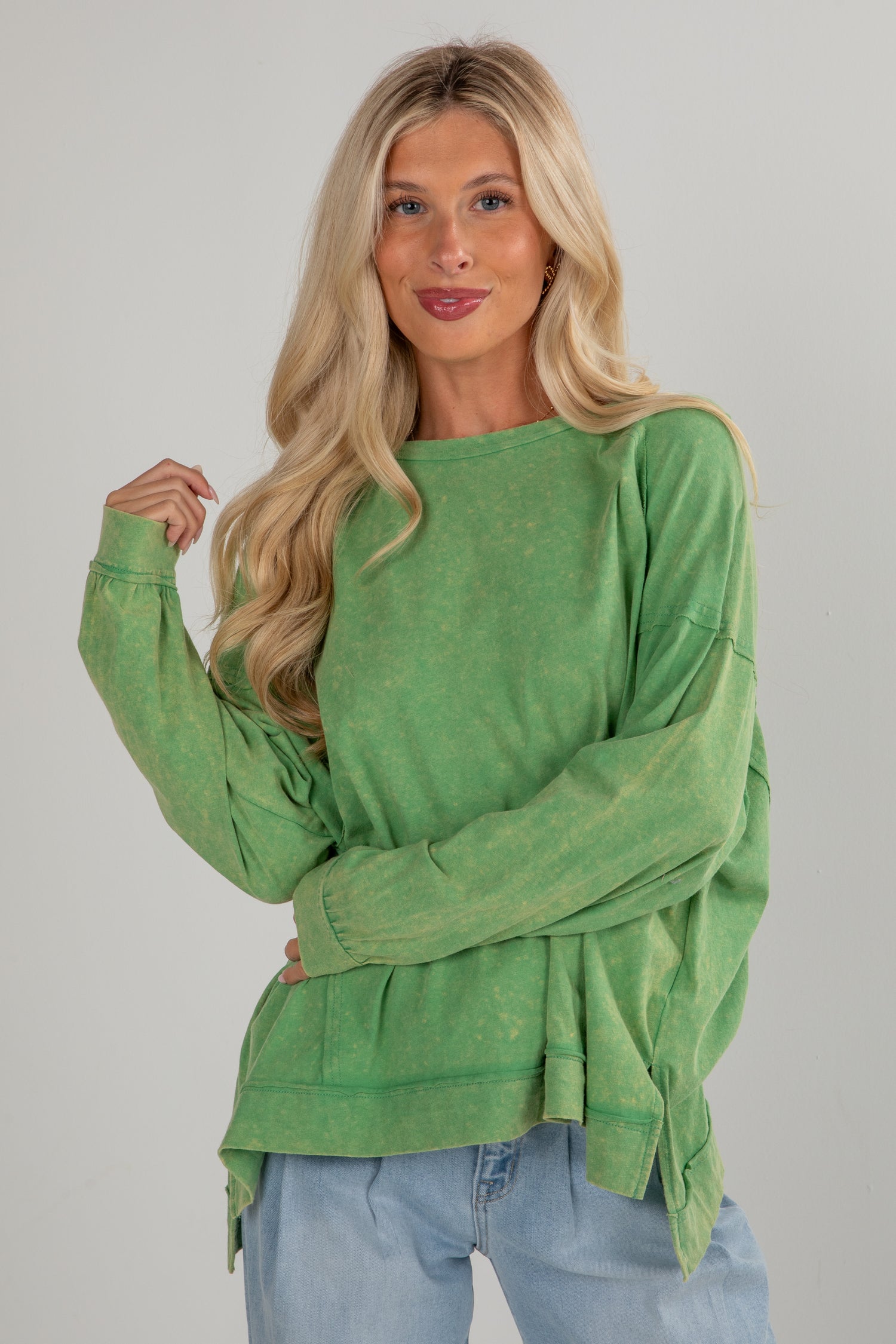 Woman wearing a green long-sleeve top and blue jeans against a gray background