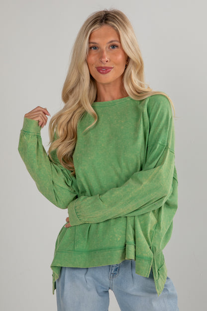 Woman wearing a green long-sleeve top and blue jeans against a gray background