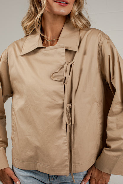 A woman wearing a beige long-sleeve utility-style jacket with an asymmetrical collar and front tie detail, paired with light blue jeans against a plain light gray background.