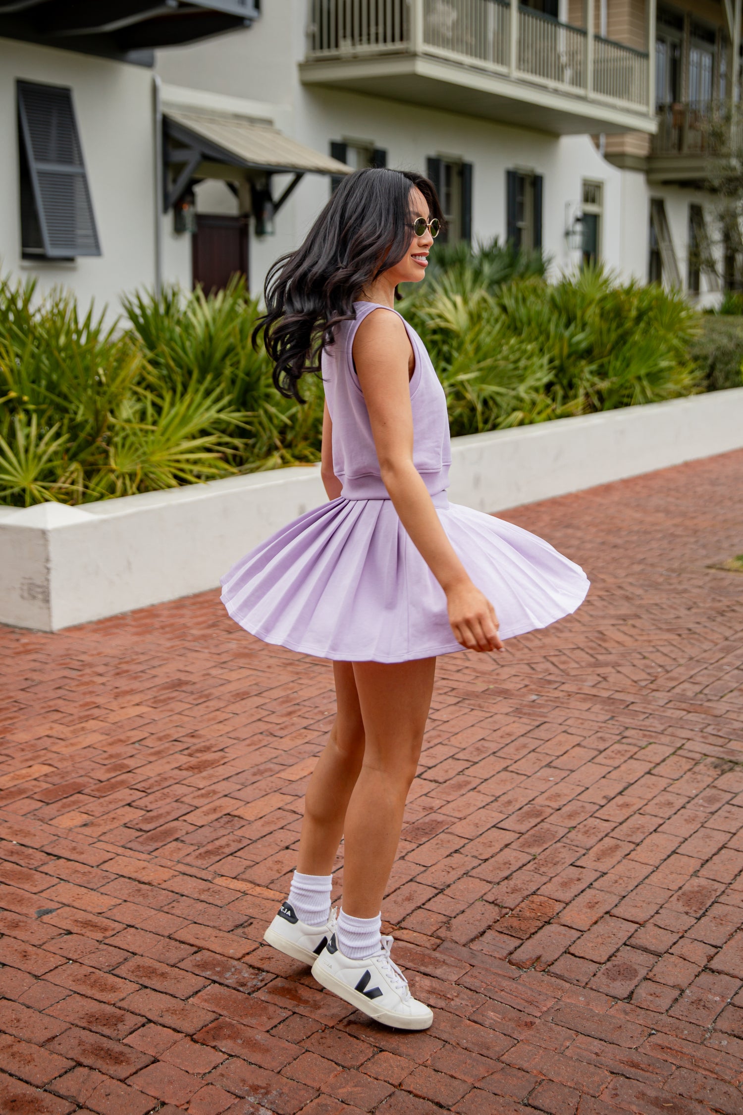 Woman in a purple dress standing on a brick path with greenery and a building in the background