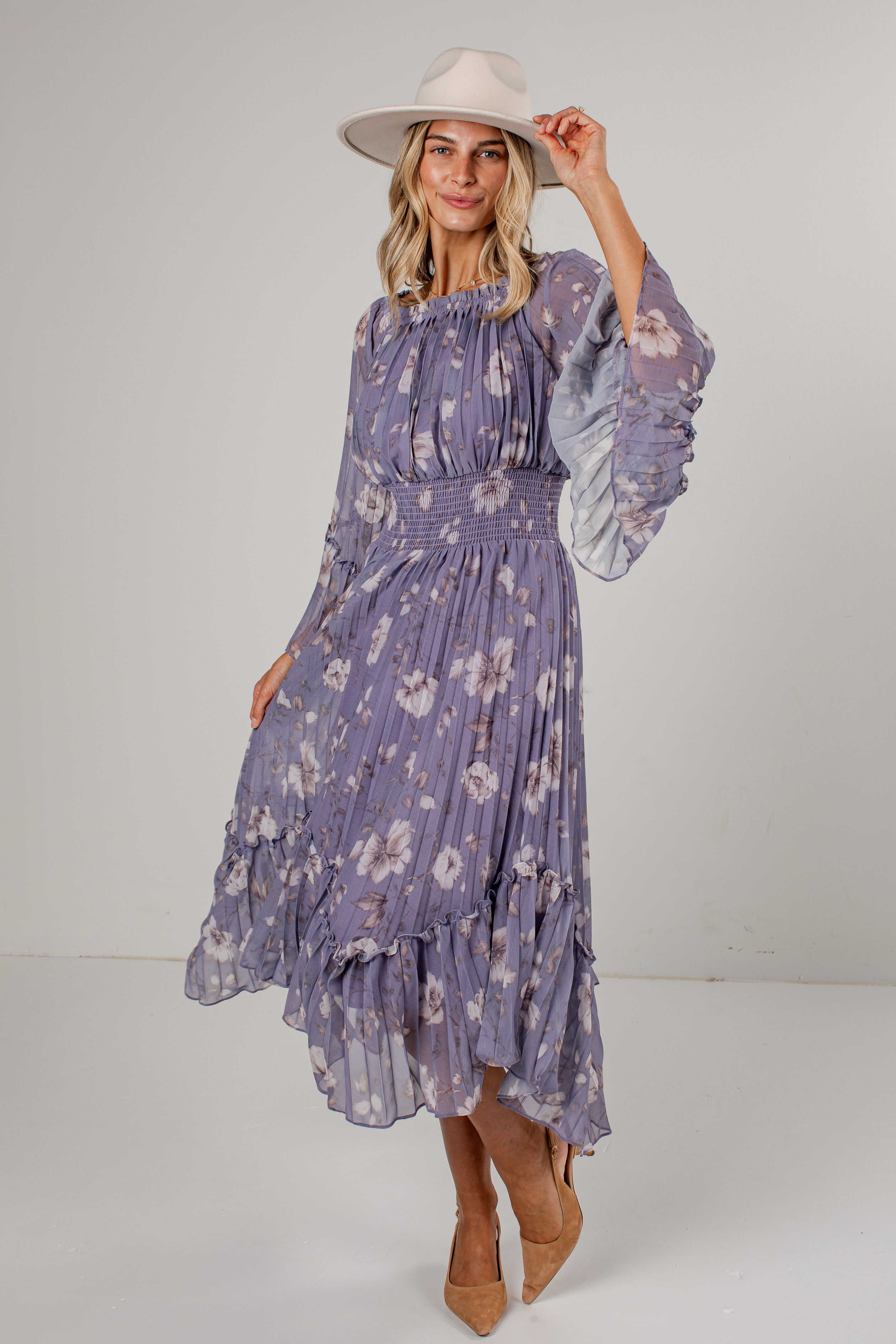 A woman modeling a lavender floral midi dress with long ruffled sleeves and a smocked waist, paired with a wide-brimmed cream hat and tan heels, against a light gray studio background.