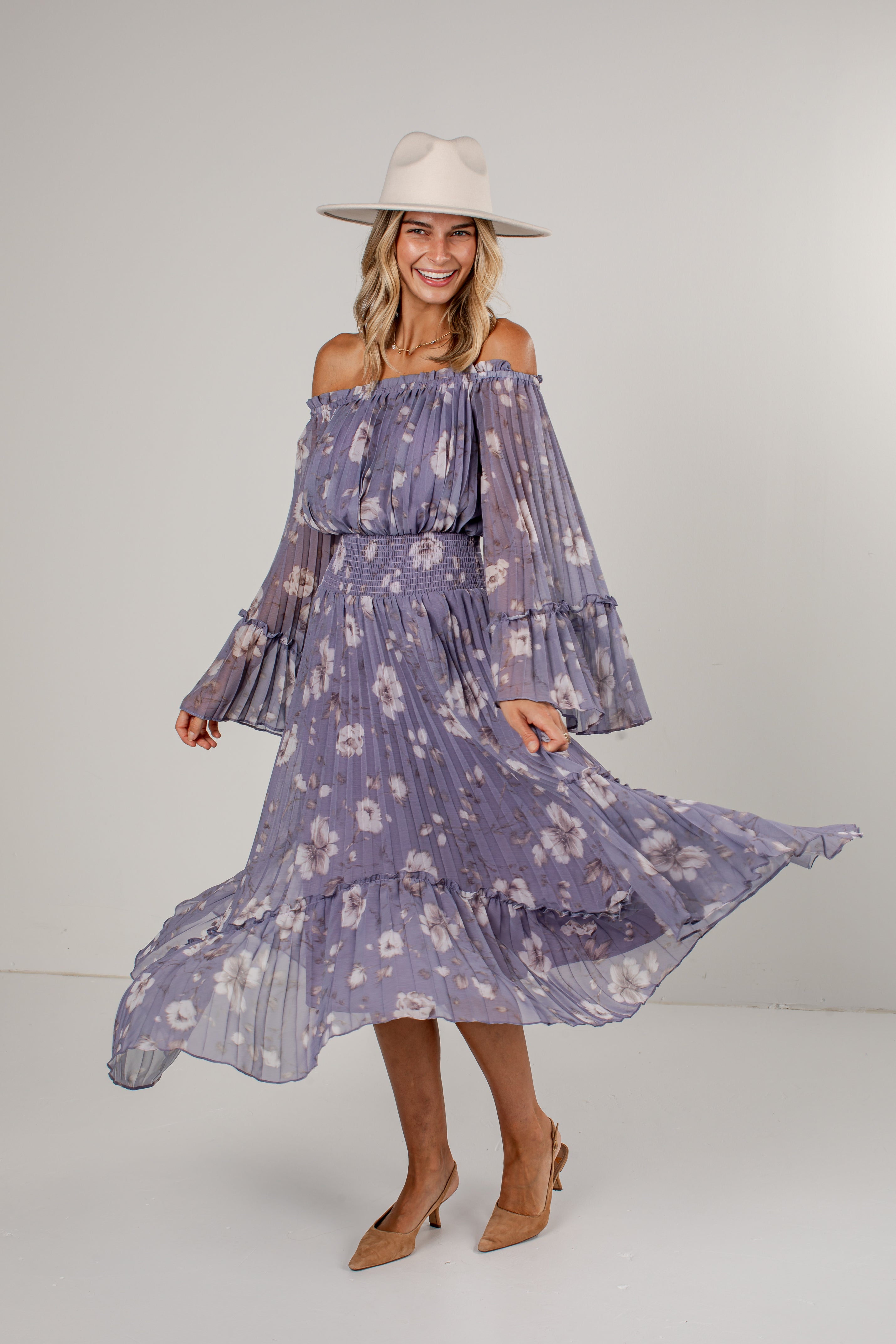 A woman modeling an off-the-shoulder midi dress with a purple and white floral print, featuring tiered long sleeves and a pleated skirt. She is wearing a beige wide-brim hat and beige heels, standing against a neutral light gray studio background.