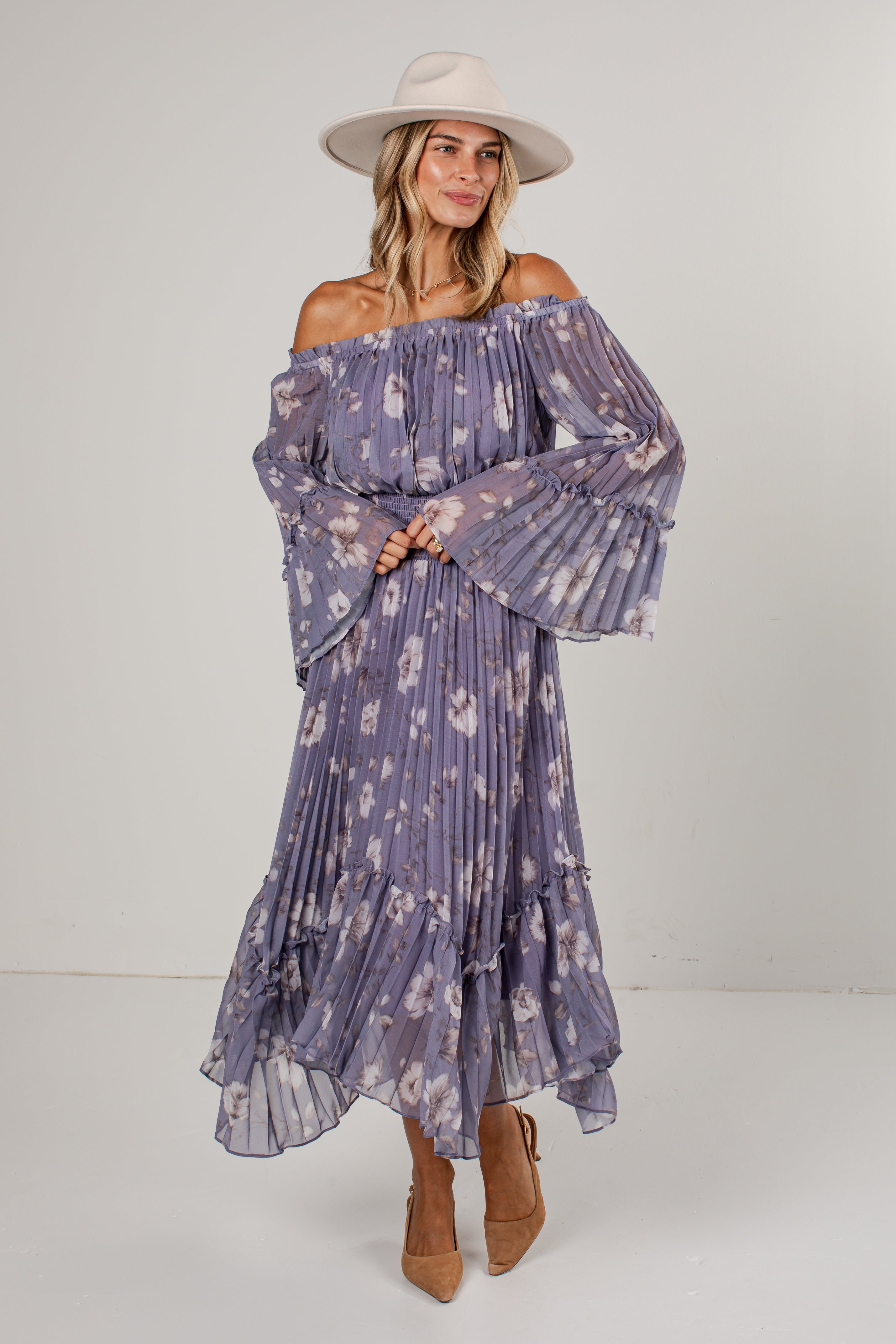 A woman modeling an off-the-shoulder, long-sleeve maxi dress with a purple and white floral print, paired with a beige wide-brim hat and tan ankle boots, against a plain light gray studio background.