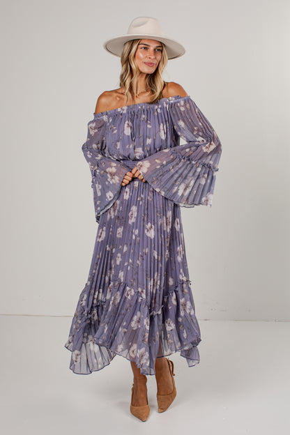 A woman modeling an off-the-shoulder, long-sleeve maxi dress with a purple and white floral print, paired with a beige wide-brim hat and tan ankle boots, against a plain light gray studio background.
