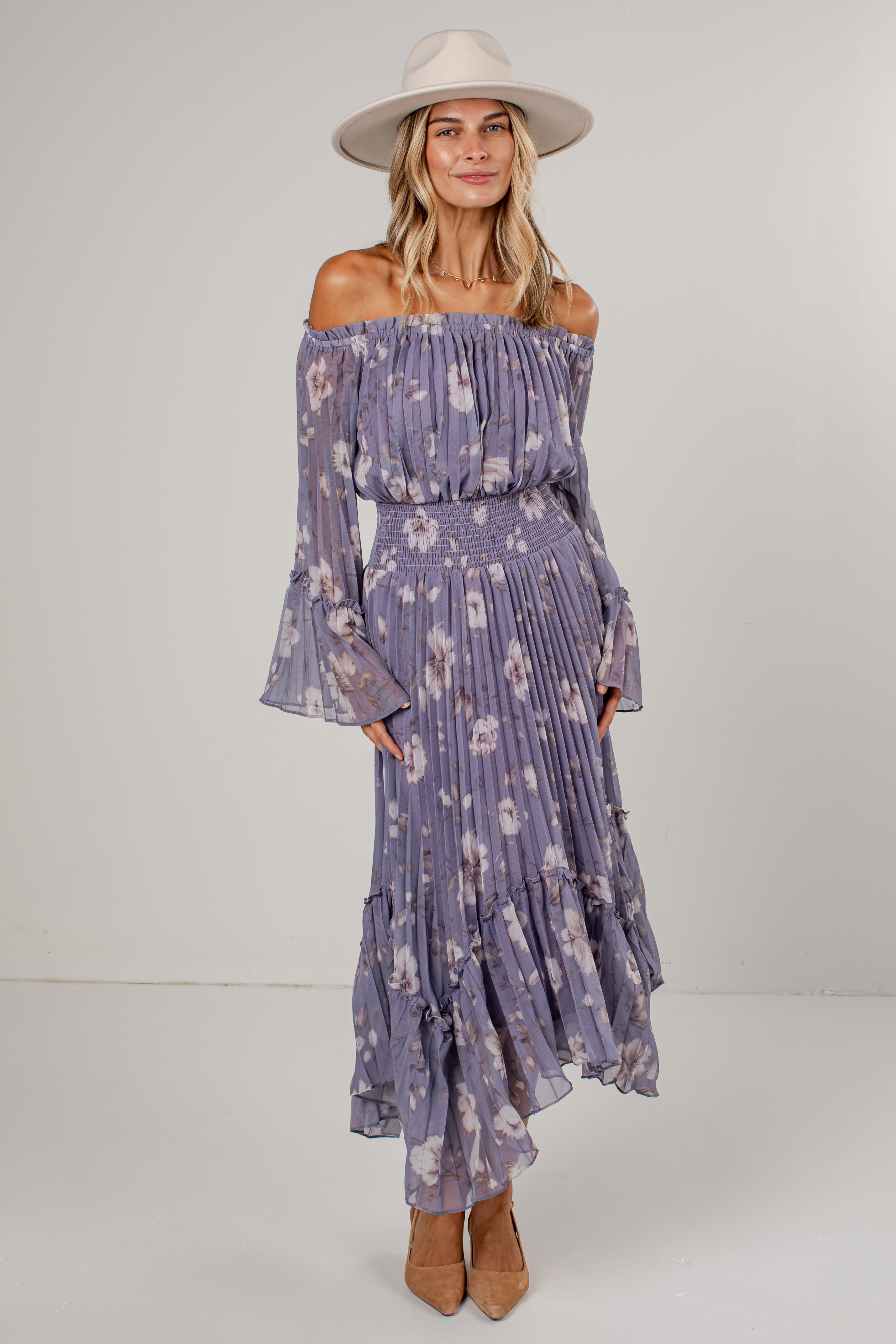 A woman modeling an off-the-shoulder midi dress in a dusty purple floral print, paired with a wide-brimmed cream hat and tan heels, against a plain light gray studio background.