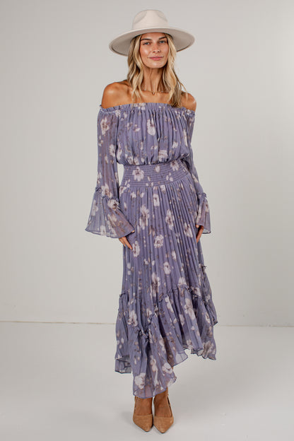 A woman modeling an off-the-shoulder midi dress in a dusty purple floral print, paired with a wide-brimmed cream hat and tan heels, against a plain light gray studio background.
