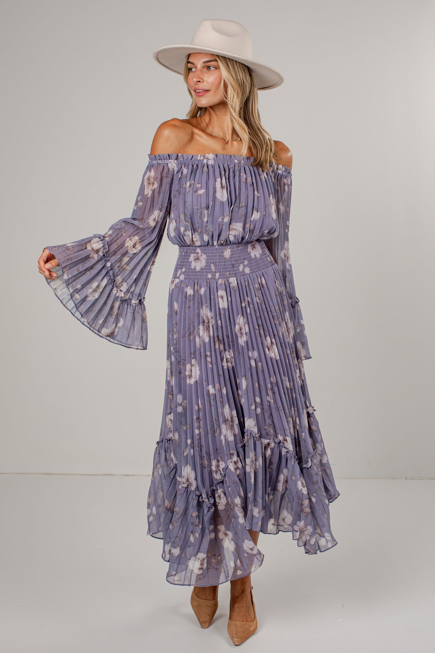 A woman modeling an off-the-shoulder midi dress with a purple and white floral print, featuring long bell sleeves, a smocked waist, and a tiered ruffle hem. She is also wearing a beige wide-brim hat and tan ankle boots, set against a plain light gray studio background.