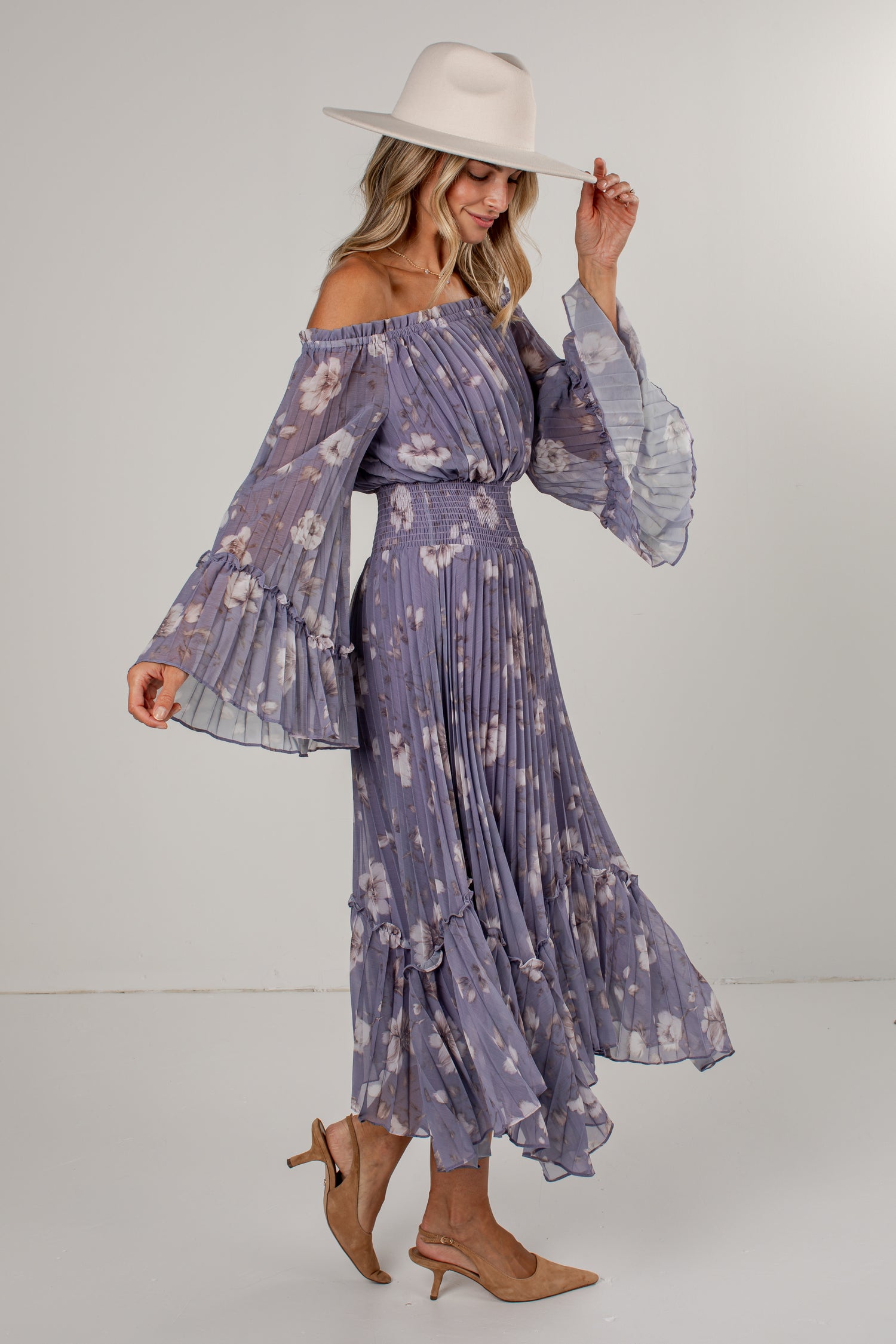 A woman modeling a lavender off-the-shoulder maxi dress with a white floral print, tiered long sleeves, and a pleated skirt, styled with a cream wide-brim hat and tan slingback heels against a light gray studio background.