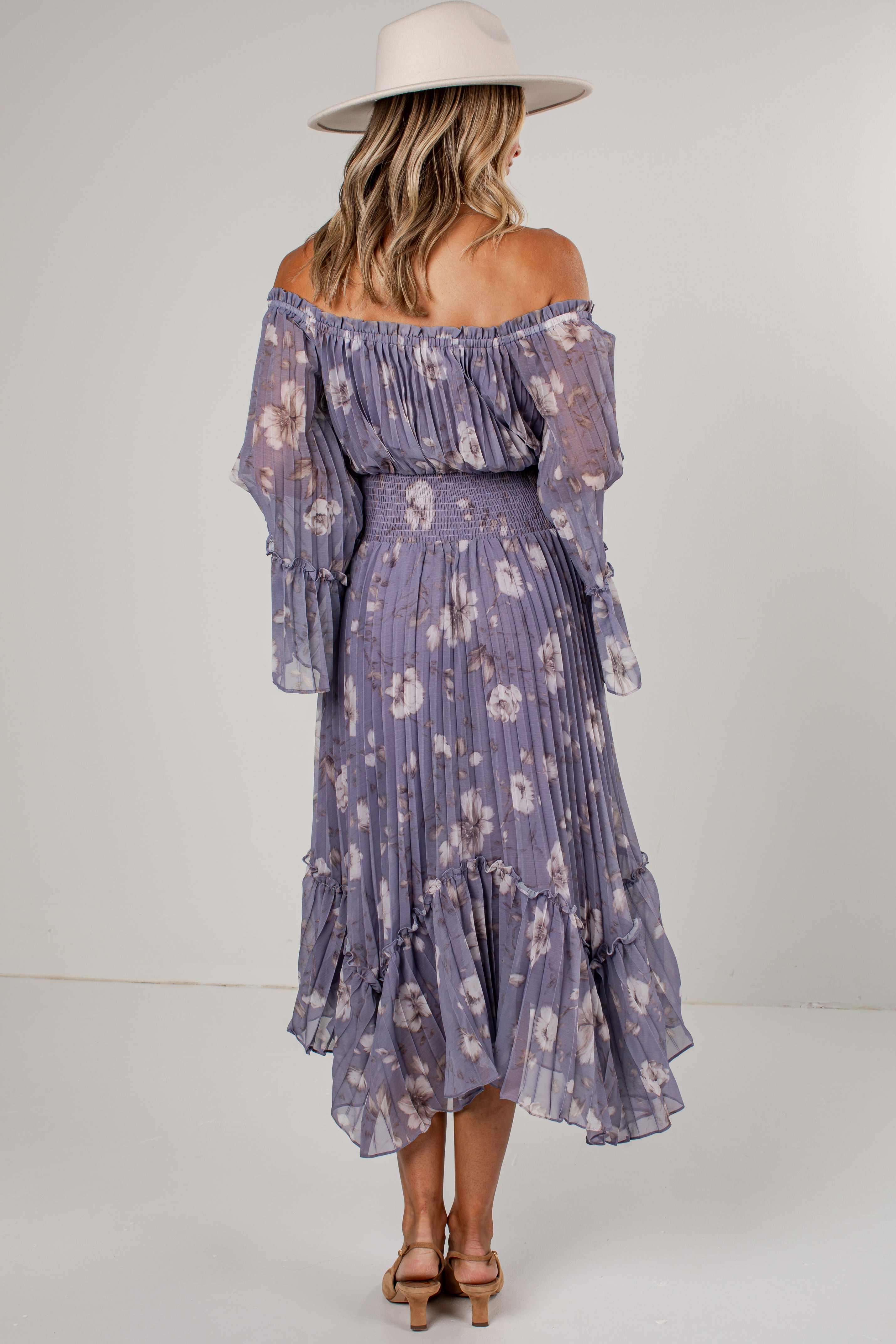 A woman shown from the back wearing a lavender off-the-shoulder midi dress with a white floral print, tiered ruffle sleeves, and a matching ruffled skirt. She is also wearing a wide-brimmed cream hat and brown strappy heels against a plain light gray studio background.
