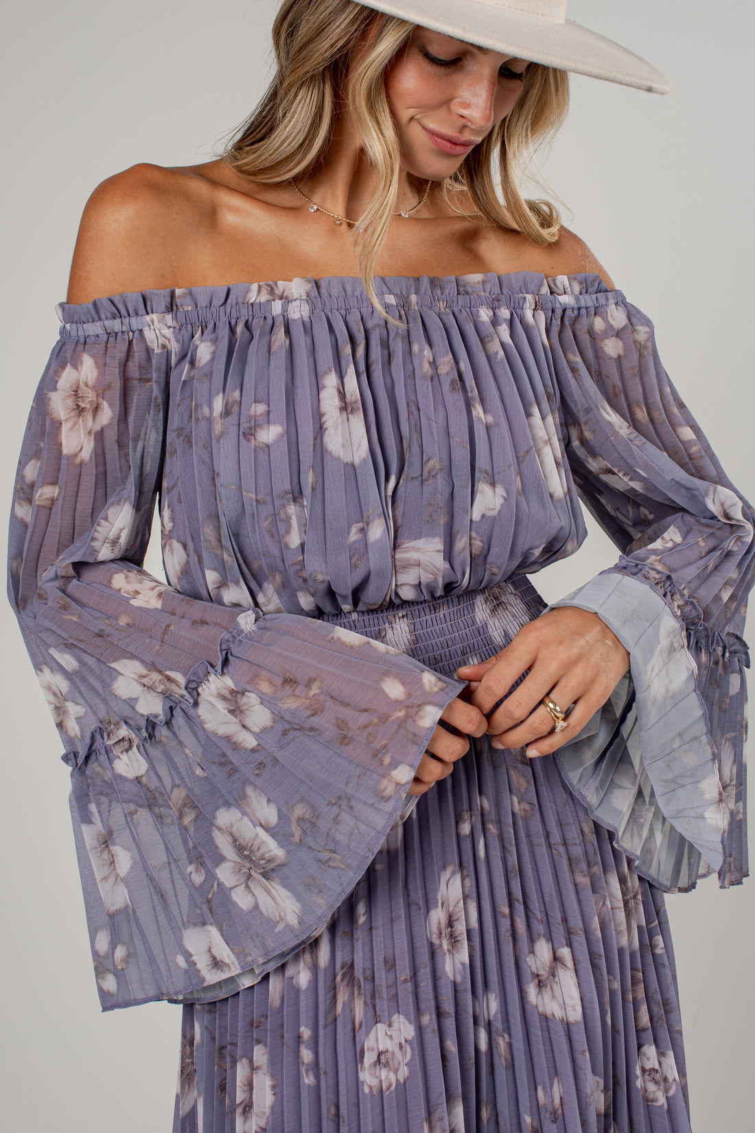 A woman wearing a lavender off-the-shoulder long-sleeve maxi dress with a white floral print, pleated fabric, and a matching pleated belt. She is also wearing a wide-brimmed cream hat, photographed against a neutral light gray background.