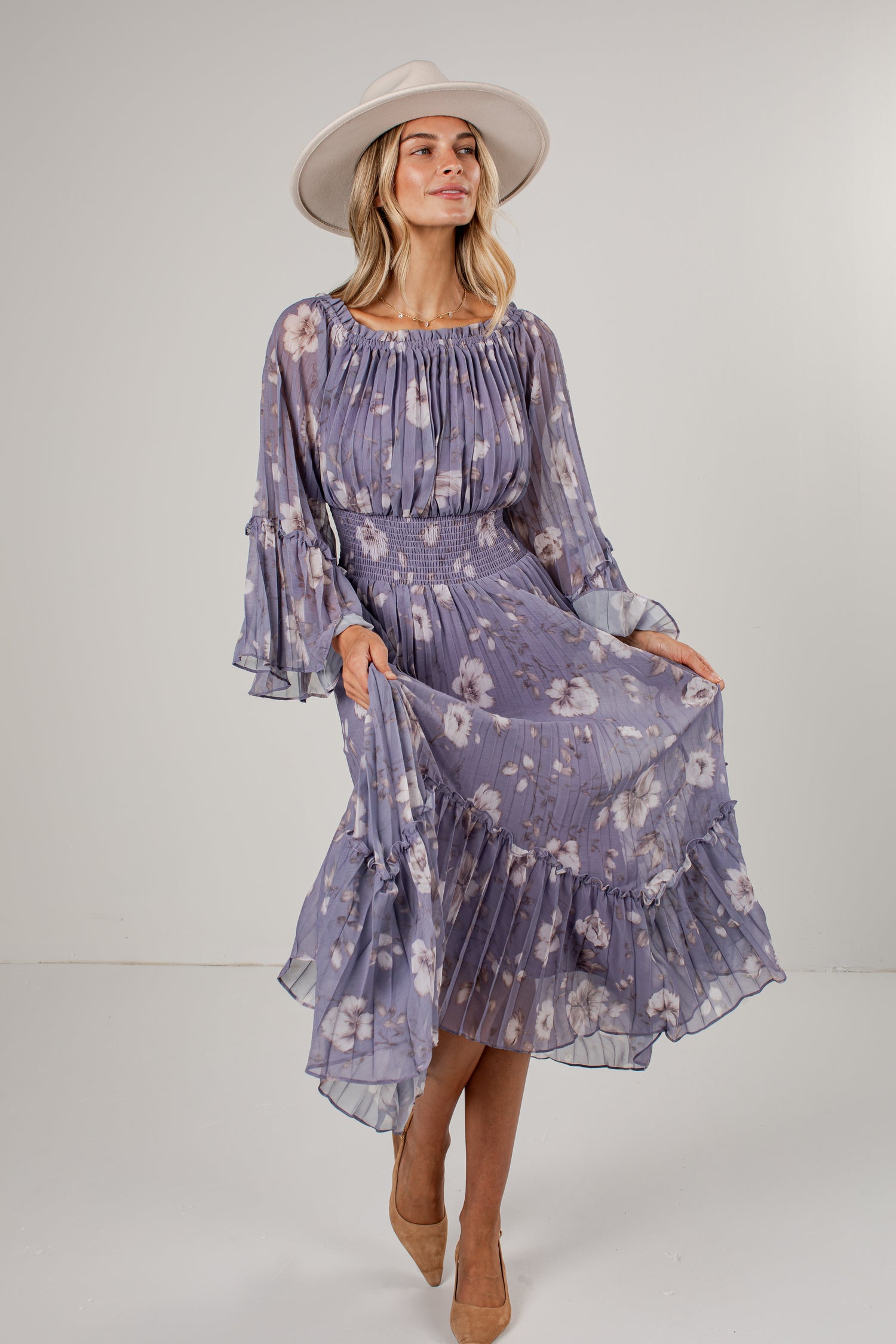 A woman modeling a lavender-purple midi dress with a white floral pattern, featuring long bell sleeves, a smocked bodice, and a tiered ruffled hem. She is wearing a wide-brimmed cream hat and tan pointed-toe heels against a plain light gray studio background.