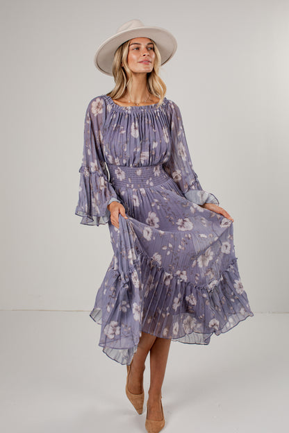 A woman modeling a lavender-purple midi dress with a white floral pattern, featuring long bell sleeves, a smocked bodice, and a tiered ruffled hem. She is wearing a wide-brimmed cream hat and tan pointed-toe heels against a plain light gray studio background.