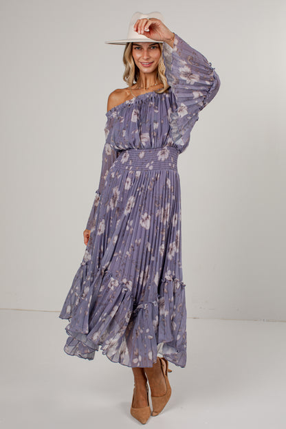 A woman modeling a long-sleeve off-the-shoulder midi dress with a purple and white floral print, paired with a white wide-brim hat and tan pointed-toe heels, set against a plain light gray studio background.