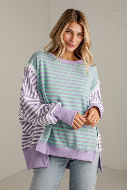 A woman modeling a lavender and mint green striped sweatshirt with solid lavender sleeves and hem, paired with light-wash jeans against a neutral indoor wall.