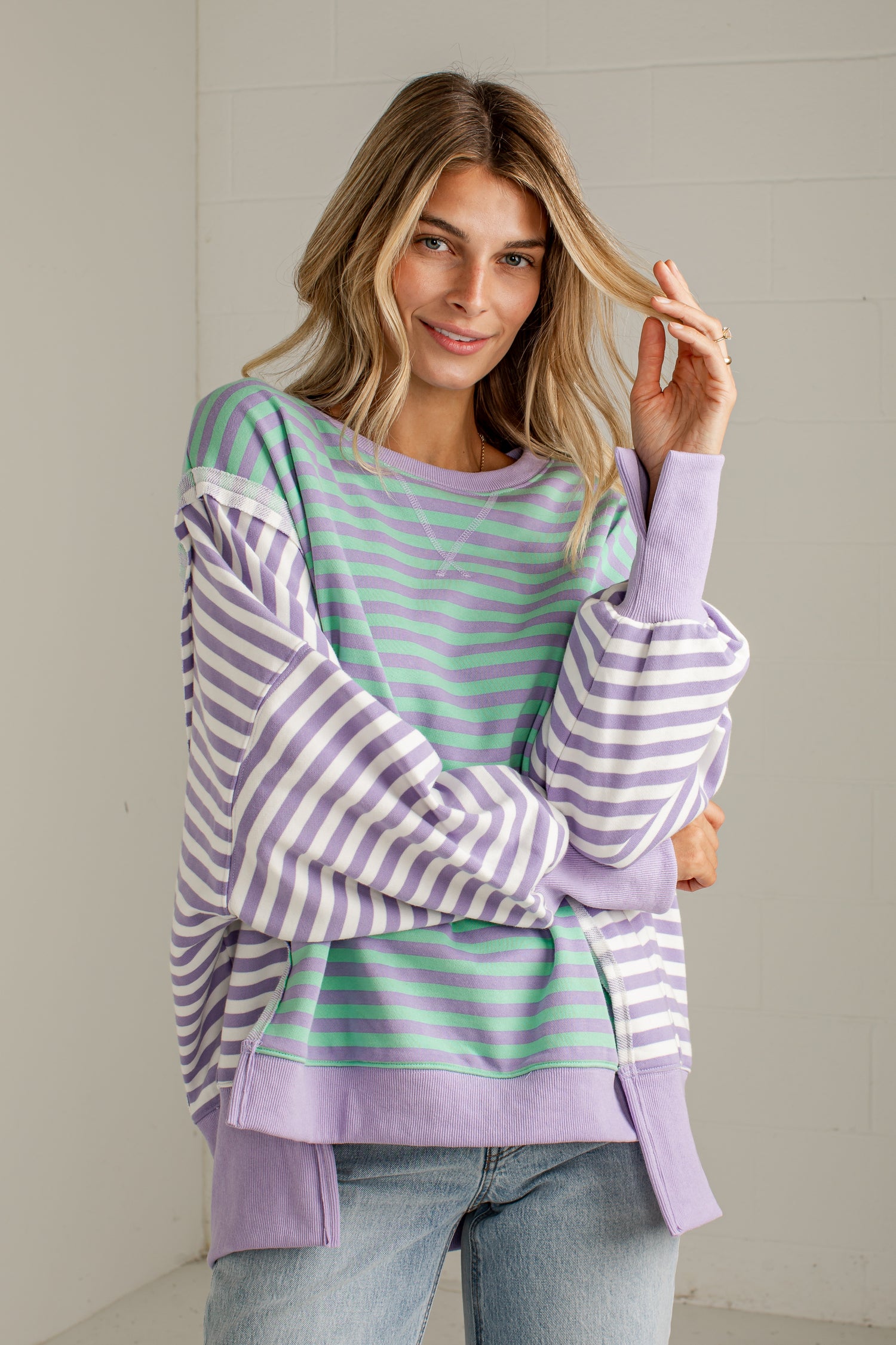 A woman wearing a lavender and mint green striped sweatshirt with balloon sleeves, paired with light blue jeans, standing against a light gray brick wall.