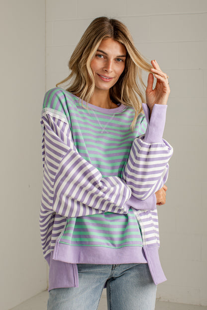 A woman wearing a lavender and mint green striped sweatshirt with balloon sleeves, paired with light blue jeans, standing against a light gray brick wall.