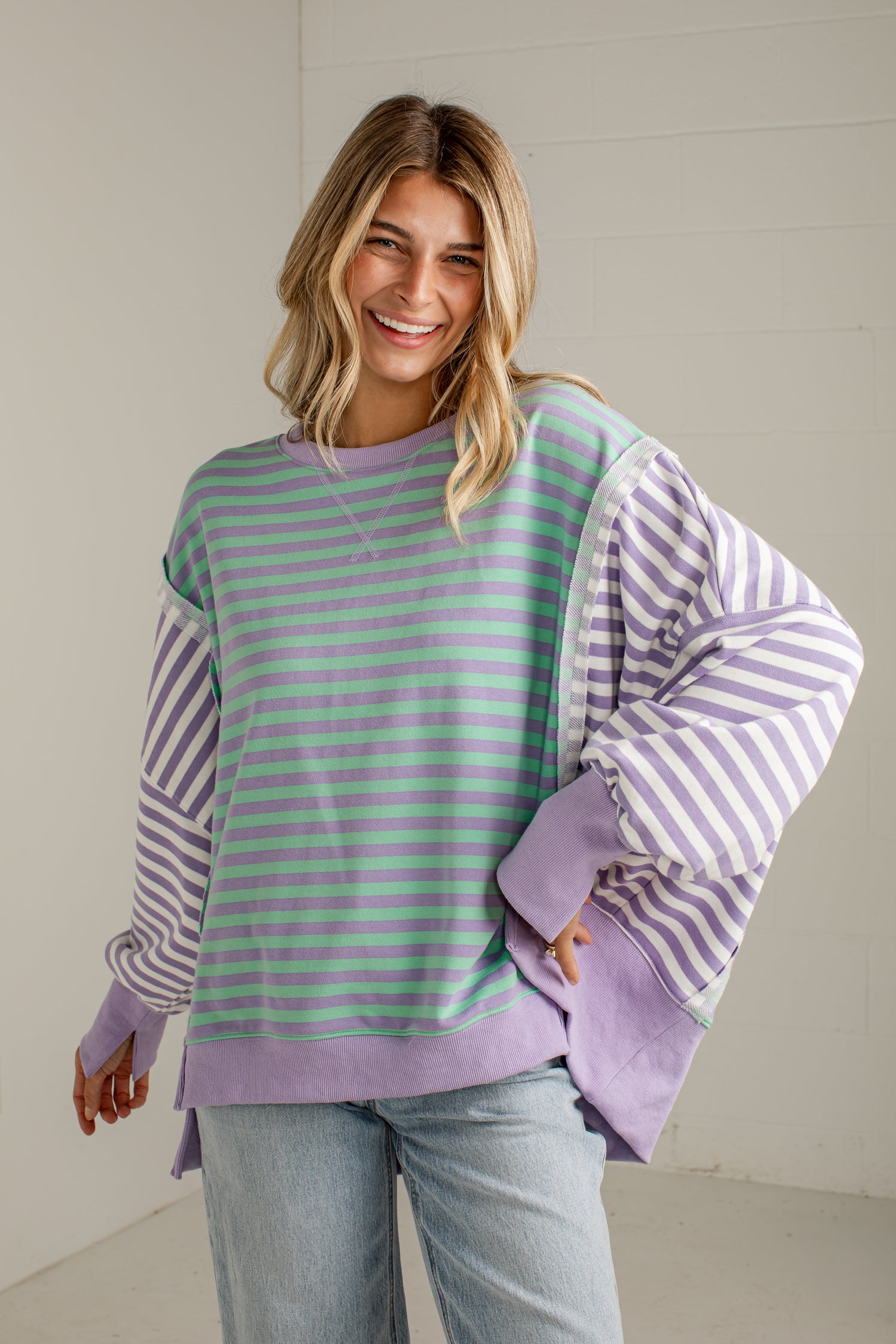 A woman wearing a lavender and mint green striped sweatshirt with solid lavender cuffs and hem, paired with light blue jeans, standing against a white paneled wall.