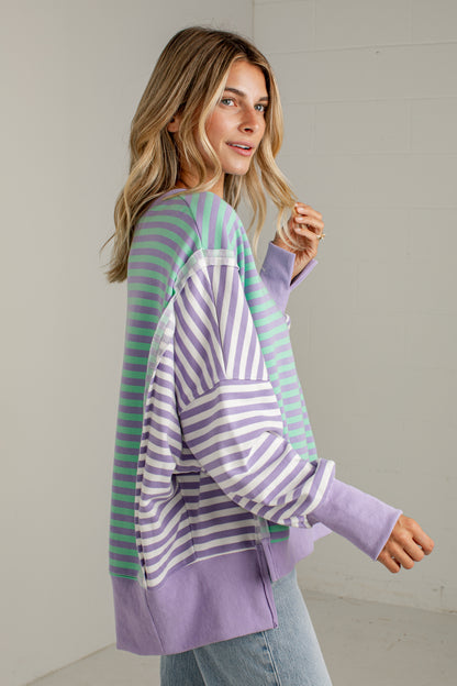 A woman wearing a lavender and mint green striped long-sleeve top, paired with light blue jeans, standing against a neutral off-white wall.