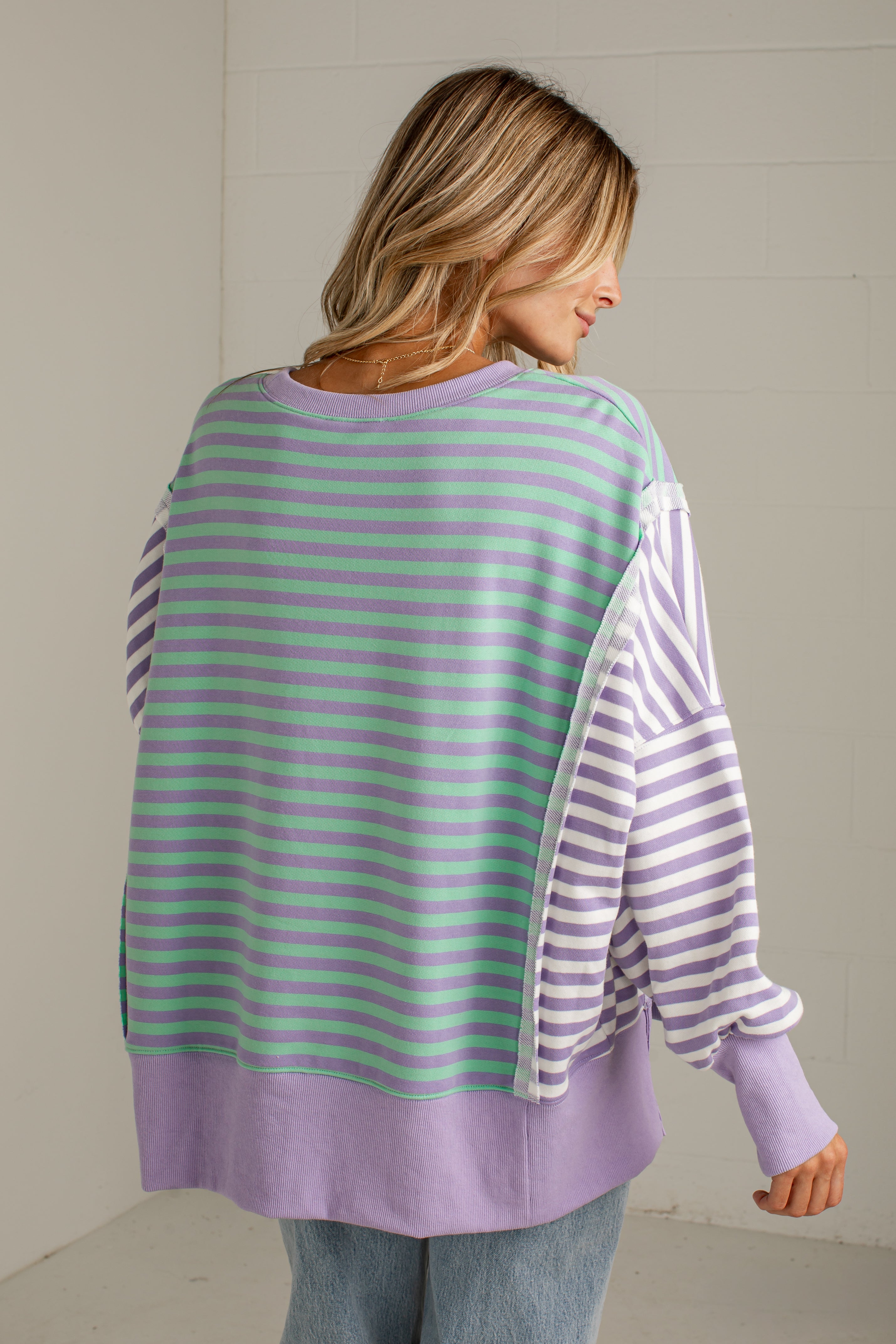A woman shown from the back wearing a lavender and mint green striped long-sleeve top with solid lavender trim, paired with light blue jeans, against a light-colored wall.