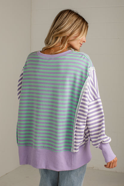 A woman shown from the back wearing a lavender and mint green striped long-sleeve top with solid lavender trim, paired with light blue jeans, against a light-colored wall.