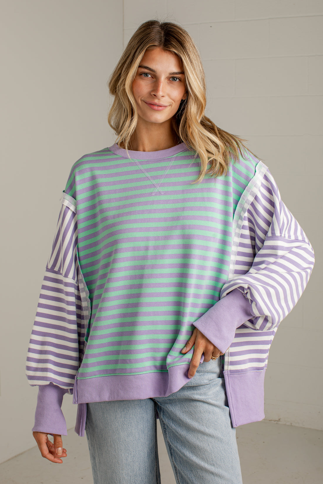 A woman wearing a lavender and mint green striped sweatshirt with solid lavender balloon sleeves, paired with light-wash jeans, standing against a neutral light gray wall.
