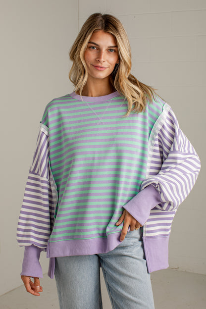 A woman wearing a lavender and mint green striped sweatshirt with solid lavender balloon sleeves, paired with light-wash jeans, standing against a neutral light gray wall.