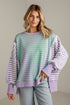 A woman wearing a lavender and mint green striped sweatshirt with solid lavender balloon sleeves, paired with light-wash jeans, standing against a neutral light gray wall.